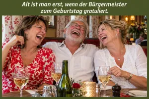 Zwei Frauen und ein älterer Mann lachen herzlich am Restauranttisch mit Weingläsern, Text: Alt ist man erst, wenn der Bürgermeister zum Geburtstag gratuliert.