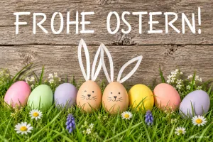 Reihe bunter Ostereier im Gras, zwei davon mit Hasenohren und Gesichtern, Text: Frohe Ostern!