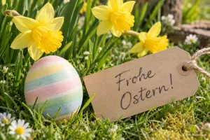 Papieranhänger neben einem gestreiften Osterei zwischen Osterglocken, Text: Frohe Ostern!