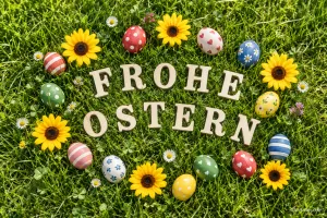 Holzbuchstaben im Gras, umgeben von bunten Ostereiern und Sonnenblumen, Text: FROHE OSTERN