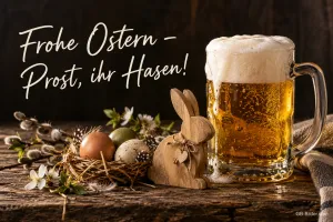 Bierkrug neben einem Holzhasen und Ostereiern, Text: Frohe Ostern – Prost, ihr Hasen!