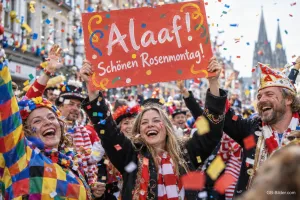 Fröhliche Menschen in Kostümen jubeln mit Schild vor dem Kölner Dom, Text: Alaaf! – Schönen Rosenmontag!